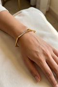 A gold geometric cuff bracelet with a row of small clear stones, worn on a person's wrist resting on a cream-colored fabric surface.