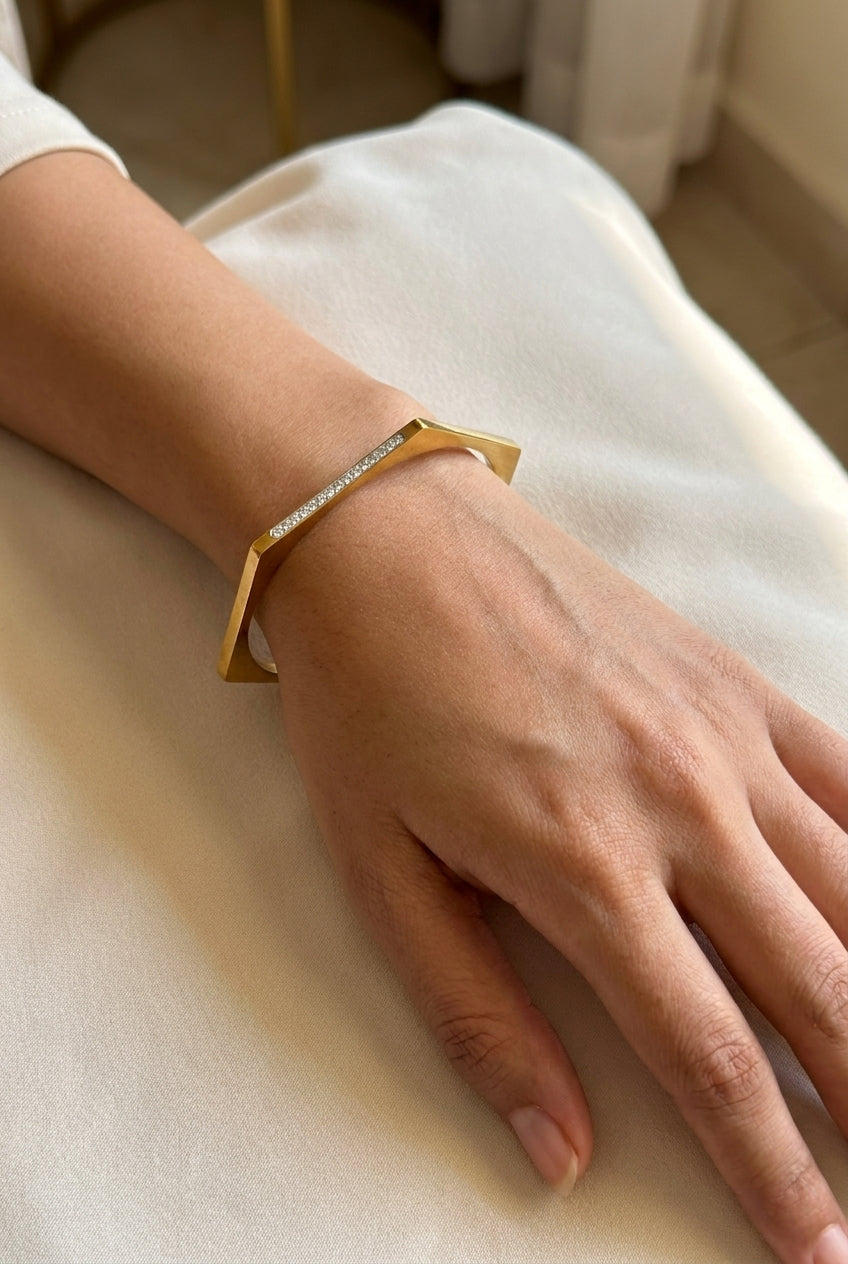 A gold geometric cuff bracelet with a row of small clear stones, worn on a person's wrist resting on a cream-colored fabric surface.