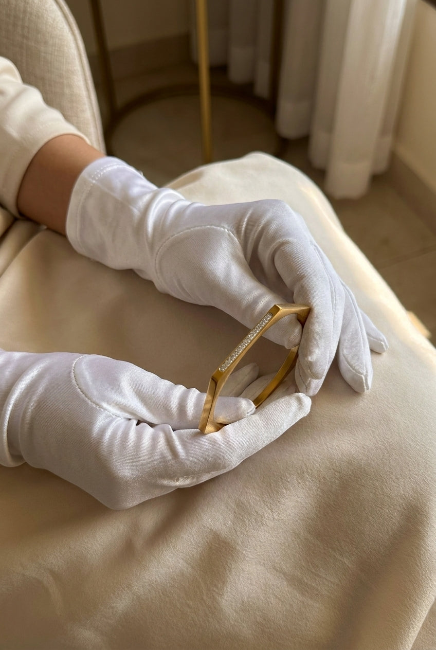 A close-up of hands wearing white gloves holding a delicate gold bangle bracelet with a row of small clear stones, set against a soft beige fabric background.