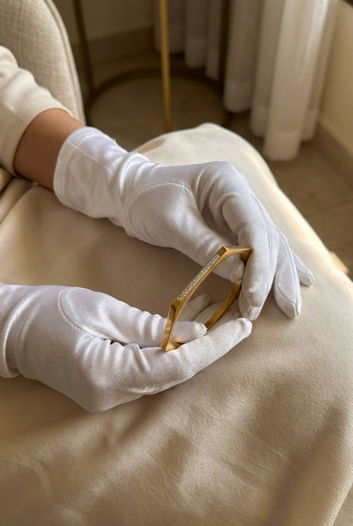 A close-up of hands wearing white gloves holding a delicate gold bangle bracelet with a row of small clear stones, set against a soft beige fabric background.