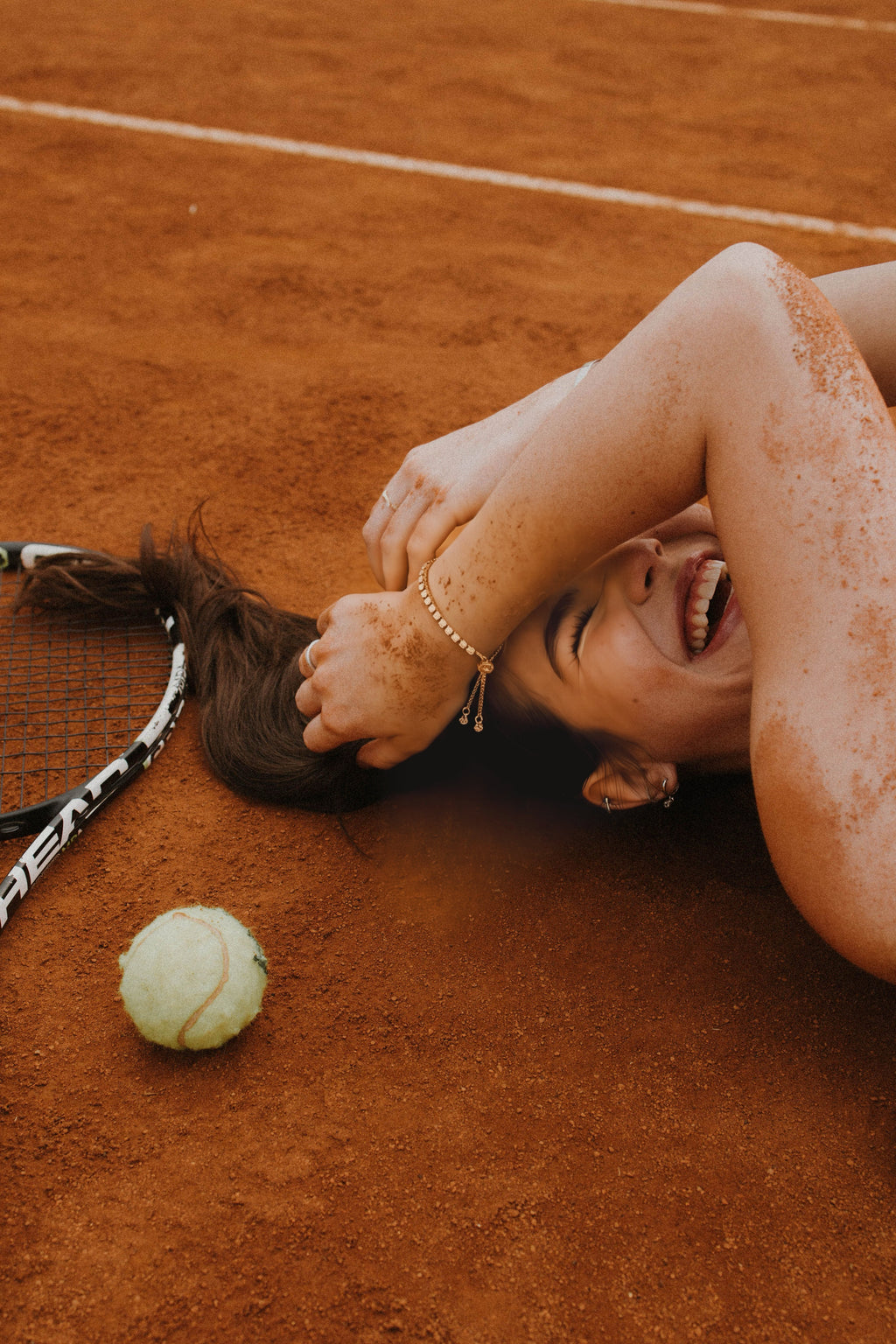 Person lying on a clay tennis court with a tennis racket and ball nearby